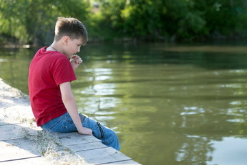 A pensive boy sits on a wooden pier, having lowered his feet in the water, and is nibbling a blade of grass, against the background of a bank overgrown with trees.