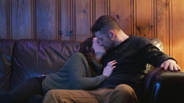 A young couple kiss as they cuddle and snuggle at night on couch, watching tv