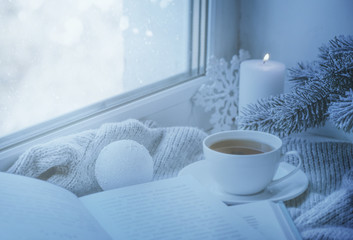 Cozy winter still life: mug of hot tea and book with warm plaid on windowsill against snow landscape from outside.