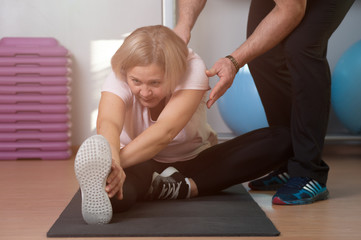Fototapeta premium An elderly woman is engaged in stretching at the gym with the help of a trainer. The concept of flexible ligaments and a healthy body in adulthood