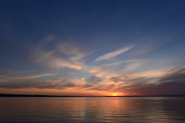 Sunlight in the clouds on the horizon under a clear blue sky at sunset over the glowing quiet surface of the lake water.