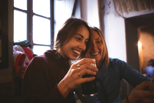 two women friends sitting in the pub with glasses of dark beer, have fun talking, laughing taking and looking photos on the smartphone