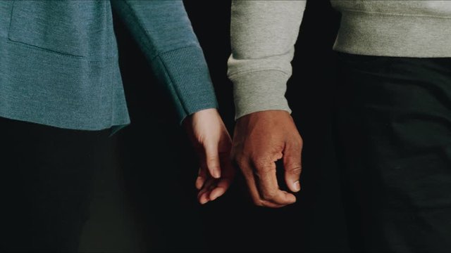 A white woman and a black man hold hands in slow motion. Their fingers interlock. They stand in front of a dark background. 