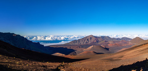 Scenic panorama of a Haleakala volcano from Keonehe'ehe'e trail overlooking cinder cones inside a caldera. Clear blue sky white puffy clouds below the horizon and rich red-brown colors of a mountain