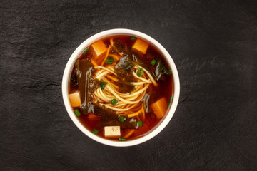 A bowl of miso shiru soup with tofu, scallions, noodles, and wakame seaweed, shot from the top on a black background with a place for text