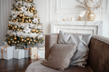 Stylish Christmas living room decor in gold and silver tones. A large beautifully decorated Christmas tree stands in the room next to the modern sofa.