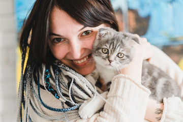 Pretty smiling young dreadlock girl with a little kitten in her hands in the cozy bedroom  