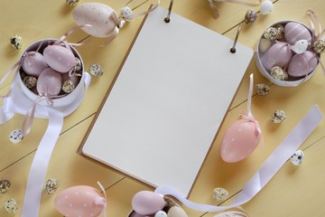  Easter Flat lay.  Easter eggs of pastel colors, blank notepad on pale yellow wooden background.Easter holiday. 