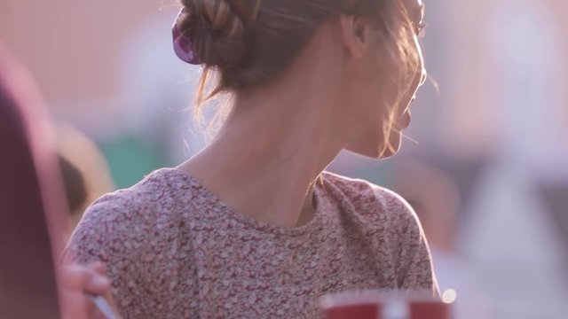 beautiful smiling woman is sitting outdoors at the table with cups of coffee, talking with someone, laughing and looking back