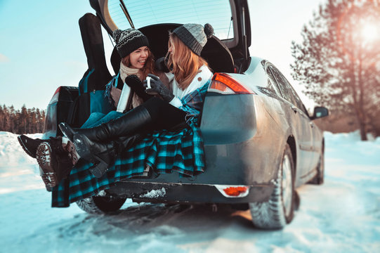 Happy friends in winter forest. Two girls sit in trunk of car drinking coffee