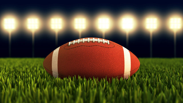 American football ball close up on field. Illuminated American soccer stadium.