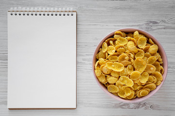 Cornflake cereal in a pink bowl for breakfast, blank notepad over white wooden background, view from above. Flat lay, overhead, top view.