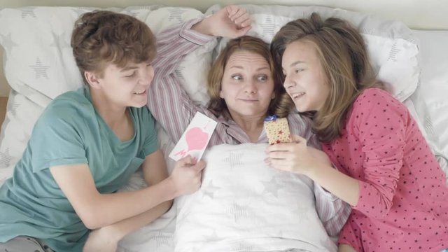 Mothers Day - Children surprise their Mum with presents in bed. Happy Family - beautiful Mother with her Son and Daughter. Girl and boy congratulates mom and gives her postcard and gift box.