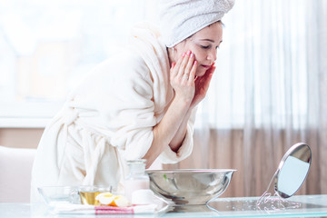 Young woman with a towel on her head washing face with water in the morning. Hygiene and care for the skin at home