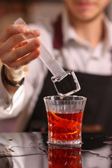 Professional bartender throwing to red cocktail glass standing on the bar counter an ice cube with splash on the blurred background