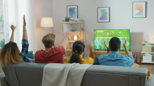 At Home Diverse Group of Sports Fans Sitting on a Couch Watching Important Soccer Match on TV, They Cheer for the Team, Celebrate Victory after Team Scoring Winning Goal. Room with Snacks and Drinks.