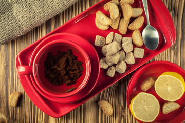 tea break. tea in a red ceramic dish on a tray with cane sugar, crackers and lemon on a wooden background. top view