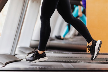 Close-up shot of athletic female legs jogging on treadmill in front of window at gym before...