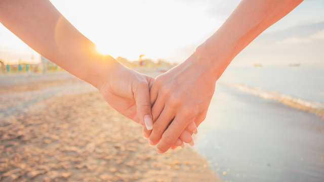 Close up of two female holding hands against sunset - two girls walking on the beach hand by hand - same-sex lesbian couple and female friendship and love, celebration of Valentine’s Day concept.