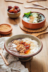 Cocoa Acia bowl with slices banana, fresh dates, candied walnuts and bee pollen topping on a brown wooden background with embroided napkins and wooden spoons