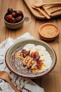 Cocoa Acia bowl with slices banana, fresh dates, candied walnuts and bee pollen topping on a brown wooden background with embroided napkins and wooden spoons