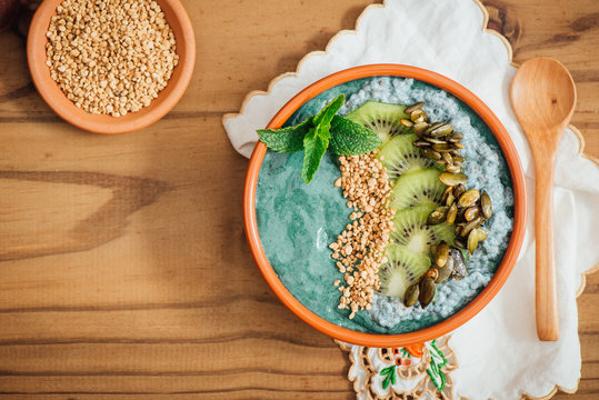 Spirulina Acia bowl with toppings of candied pumpkin seeds, kiwi fruit, bee pollen and fresh mint, on a brown wooden background