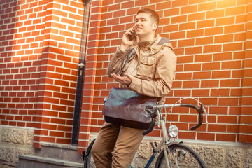 Fototapeta na wymiar Young stylish businessman with his smartphone in hand stay near red wall with sport retro bike in sunny day