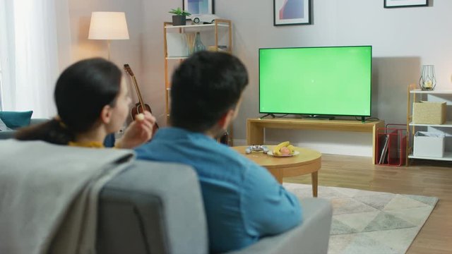 Happy Couple Sitting At Home in the Living Room Watching Green Chroma Key Screen Television, Relaxing on a Couch. Couple Room Watching Sports Game, News, TV Show or a Movie.
