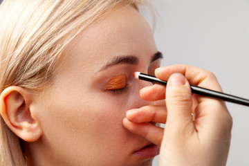 Makeup artist with a brush in his hand put on make-up dense golden shadows on the eyelids of a close-up model in a visage studio in a training lesson.