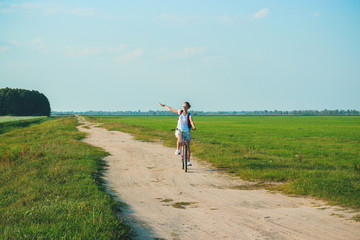 Young beautiful woman rides on a bicycle in the summertime on the green fields background and stretched out hand in the air