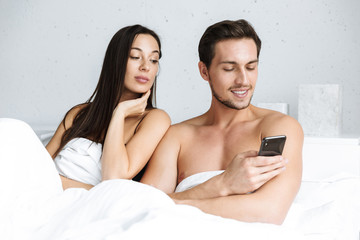 Image of young couple using cell phone together, while lying in bed at home or hotel apartment