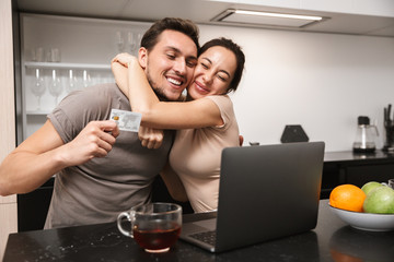 Photo of modern couple man and woman using laptop with credit card, while sitting in kitchen