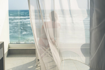 Naked beautiful female in the bikini sitting on a chair behind the transparent waving curtains on the glass balcony at the summer windy day. Sea view
