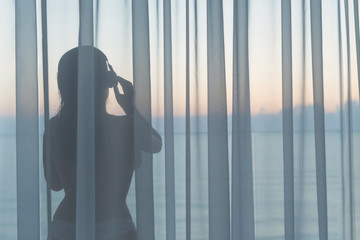 Naked beautiful woman in the headphones and dancing behind the transparent curtain on the glass balcony early morning at sunrise. Sea view.