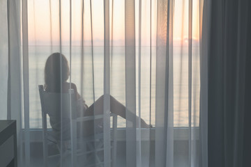 Fototapeta premium Naked beautiful female wrapped in a blanket sitting on a chair behind the transparent curtains on the glass balcony early morning at sunrise. Sea view