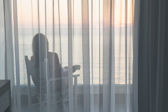 Naked beautiful female wrapped in a blanket sitting on a chair with cup of coffee behind the transparent curtains on the glass balcony early morning at sunrise. Sea view