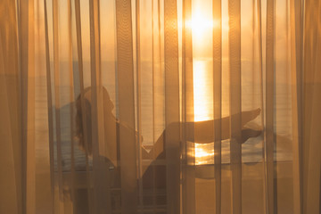 Naked beautiful girl stands behind the transparent curtain on the balcony at sunrise on the tropical resort
