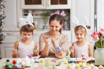 Happy young mother and her two little daughters with white rabbit's ears on their heads dye the eggs for the Easter table in the cozy light kitchen