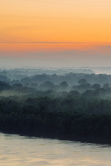 Fototapeta na wymiar Mystical view on riverbank of large island with forest under haze at early morning. Mist among layers from tree silhouettes under warm predawn sky. Morning atmospheric landscape of majestic nature.