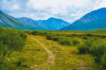 Fototapeta na wymiar Spectacular view of distant giant mountains. Footpath through valley in highland in sunny day. Hiking path. Wonderful huge mountain range under cloudy sky. Amazing green landscape of majestic nature.