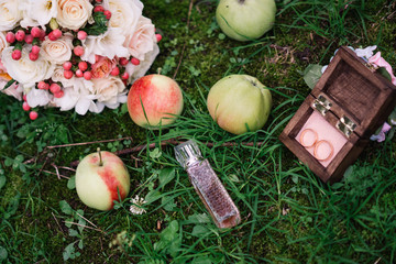 wedding rings in a wooden box in the grass with apples and perfumes with a wedding bouquet of flowers