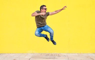 Fototapeta na wymiar Front view of a young man wearing sunglasses jumping against a yellow bright wall in a sunny day