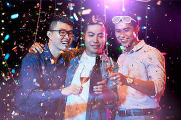 Joyful young Asian people drinking champagne under falling confetti at night party