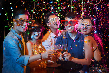 Happy excited young Vietnamese people in party glasses posing with champagne glasses under fallling...
