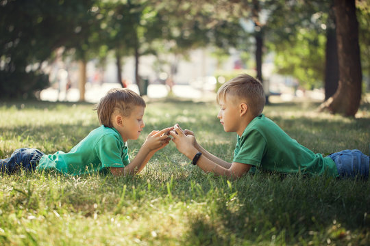 Portrait Of Two Kids Lying On Green Grass And Playing In Mobile Phone.