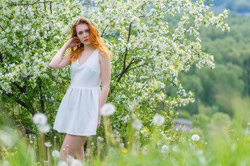 Obraz premium Beautiful and young girl standing next to a blooming Apple tree in a white dress. Red lips of the girl.