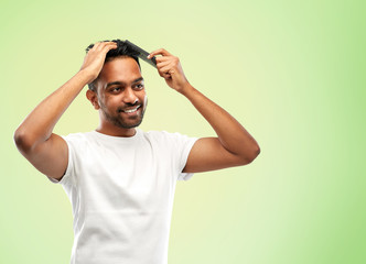 Fototapeta na wymiar grooming, hairstyling and people concept - smiling young indian man brushing hair with comb over green background