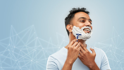 grooming and people concept - young indian man shaving beard with manual razor blade over blue background with low poly shape projection