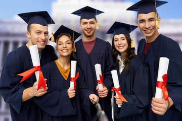 education, graduation and people concept - group of happy graduate students in mortar boards and bachelor gowns with diplomas taking picture by slfie stick over white background