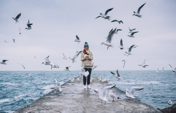 Happy young tourist woman feeds seagulls on the sea. Pretty female wearing coat, scarf and watching flying seagulls by the sea in the sky. People, travel, environment, nature concept.